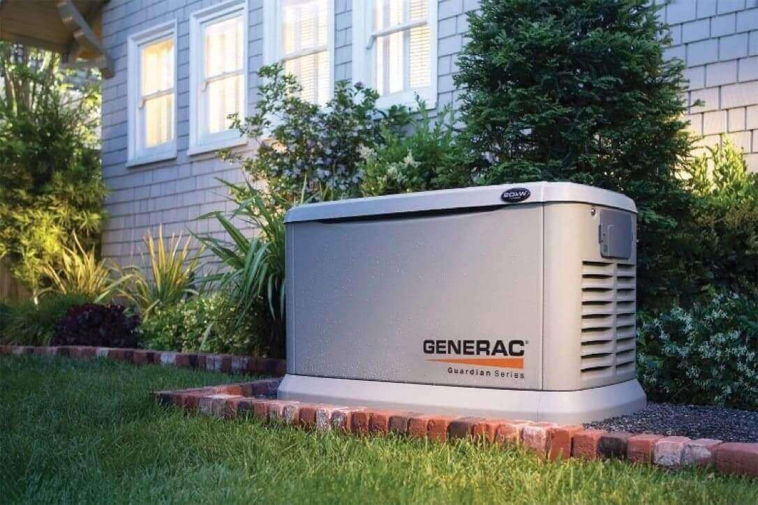 A Generac generator installed on the outside of a home. The lights are on in the home and there are lights on among the bushes between the generator and the house.